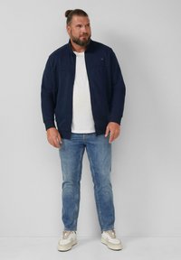 Giacca blu navy con zip frontale e colletto alto, abbinata a una t-shirt bianca, jeans blu e scarpe da ginnastica chiare, in piedi contro uno sfondo neutro.
