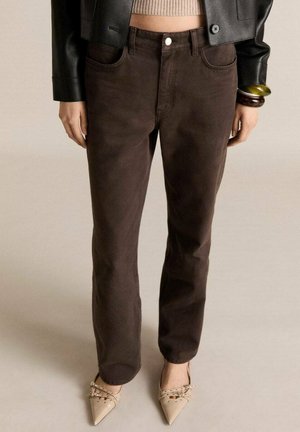 Femme portant un pantalon marron foncé à jambes droites, des chaussures beiges pointues avec des brides, un pull court beige, une veste en cuir noire et des bracelets au poignet.