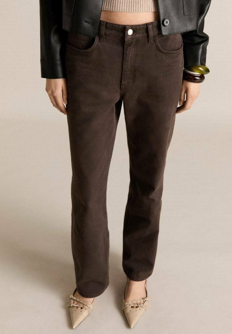 Femme portant un pantalon marron foncé à jambes droites, des chaussures beiges pointues avec des brides, un pull court beige, une veste en cuir noire et des bracelets au poignet.