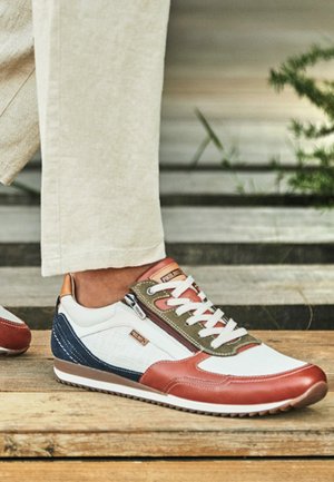 Sneaker casual con pannelli bianchi, marroni, blu navy e oliva, lacci bianchi e cerniera, indossata con pantaloni beige su superficie in legno.