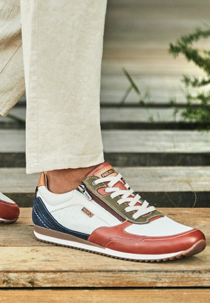 Zapatilla deportiva informal con paneles blancos, marrones, azul marino y oliva, cordones blancos y cremallera, usada con pantalones beige sobre una superficie de madera.