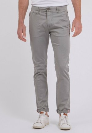 Homme portant un pantalon gris clair ajusté avec les revers retroussés et des baskets blanches, debout devant un fond uni.