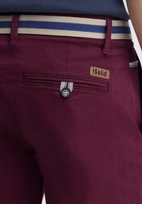 Pantalons bordeaux avec une ceinture rayée, dotés d'une poche arrière avec fermeture à bouton et d'une étiquette de marque en cuir marron attachée.