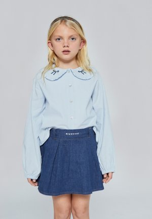 Camisa de manga larga azul claro con cuello con volantes y lazos azul marino, combinada con una falda plisada de mezclilla oscura que presenta una cinturilla con la marca.