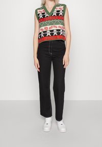 Grünes gestricktes ärmelloses Oberteil mit festlichem Muster; enthält den Text „Ho Ho Ho“, Lebkuchenmänner und Zuckerstangen. Kombiniert mit schwarzen hochtaillierten Jeans.