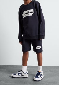 Navyblauwe sweatshirts en bijpassende shorts, gemaakt van katoenmix materiaal, met een logo patch. Afgewerkt met witte en navy sneakers en crew sokken.