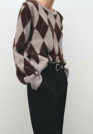 Person iført en brun og grå argyle sweater stoppet ind i sorte bukser med høj talje og et sort bælte, stående mod en hvid tekstureret væg.