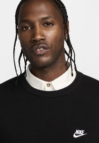 Μαύρος πουλόβερ με ρινωτή υφή, με λευκό λογότυπο Nike στο στήθος. Φοριέται πάνω από ένα λευκό πουκάμισο με γιακά και λεπτομέρεια κουμπιού.