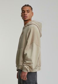 Hoodie beige surdimensionné avec une poche avant, des poignets côtelés et une coupe décontractée. Présente un imprimé logo discret sur la poitrine.