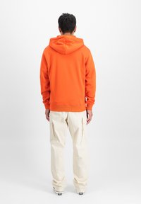 Sudadera naranja con bolsillo canguro, puños y dobladillo acanalados; combinada con pantalones cargo beige de corte holgado que cuentan con bolsillos laterales.