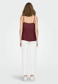 Camiseta burdeos con tirantes finos, textura suave y corte suelto, combinada con pantalones anchos blancos y zapatillas blancas, vista desde la espalda.