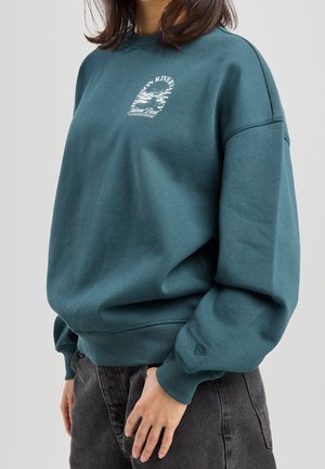 Persoon die een teal gekleurde ronde hals trui met witte grafische tekst en donkergrijze spijkerbroek draagt, staand met één hand op de heup.