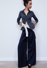 Gray button-up shirt with long sleeves and an open collar, paired with dark blue pinstripe wide-leg pants. White sweater tied at waist.