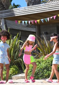 Maillots de bain pour enfants de couleur vive : bikini néon rose et jaune, chapeau de soleil, tenues en denim, et sandales roses. Cadre tropical avec de la verdure.