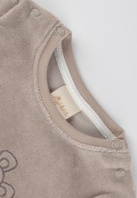 Beiger Baby-Body mit weicher Textur, Metall-Druckknöpfen an der Schulter und einem gewebten Etikett am Ausschnitt. Verfügt über ein dezentes gesticktes Design.
