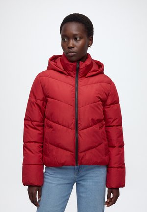 Femme portant une doudoune matelassée rouge avec capuche et un jean bleu clair, regardant vers sa droite sur un fond blanc uni.
