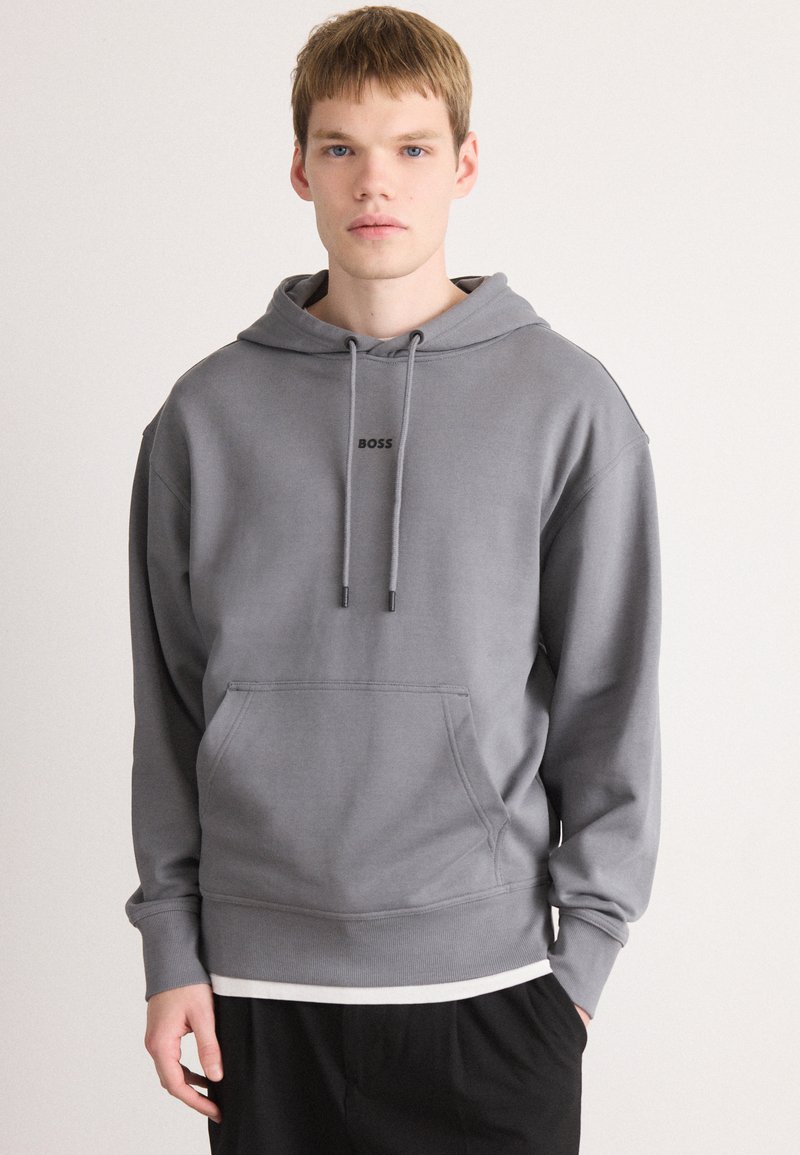 Jonge man met kort haar, gekleed in een grijze hoodie met de tekst "BOSS", zwarte broek en een witte shirt eronder, staand tegen een witte achtergrond.