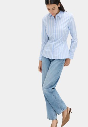 Femme portant une chemise boutonnée à rayures bleu clair, un jean bleu clair et des chaussures à talons marron avec bride arrière, debout devant un fond uni.