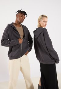 Siva zip-up dukserica s kapuljačom s prednjim džepovima; nose je dva modela, jedan okrenut prema naprijed, drugi okrenut bočno, složeno preko svijetlosmeđeg majice.