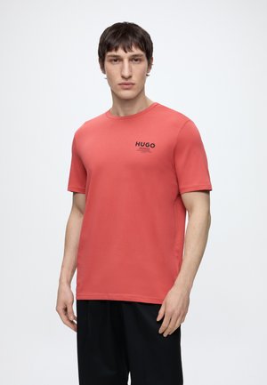 Jeune homme portant un T-shirt corail à manches courtes avec le logo noir « HUGO » sur la poitrine et un pantalon noir ample, debout sur fond blanc.