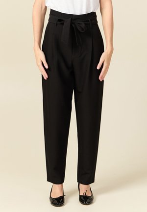 Donna che indossa pantaloni neri a vita alta con gamba affusolata, dotati di una cintura di tessuto annodata a fiocco, abbinati a ballerine nere in vernice.