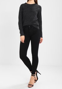 Dunkelgrauer Strickpullover mit langen Ärmeln, kombiniert mit eng anliegenden schwarzen Jeans und schwarzen Riemchen-Pumps. Einfaches, elegantes Design ohne sichtbare Muster.