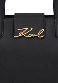 Sac à main en cuir noir avec un design cousu, une forme structurée et un logo "Karl" doré sur le rabat avant. Texture lisse.