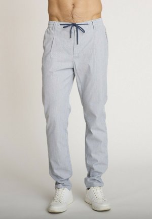Homme portant un pantalon bleu clair à cordon avec des rayures subtiles et des baskets blanches, debout sur un fond uni.