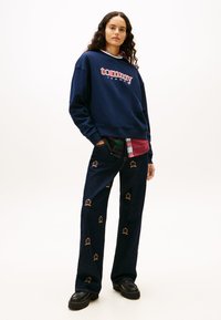 Ναυτικό φλις φούτερ με το λογότυπο "tommy", φορεμένο πάνω από κόκκινο και πράσινο πουκαμισό με καρό μοτίβο· συνδυασμένο με σκούρα τζιν παντελόνια διακοσμημένα με κεντημένα στέμματα.