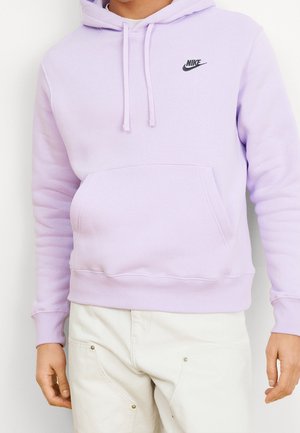 Persona vestida con una sudadera Nike de color púrpura claro con bolsillo canguro y pantalones blancos, de pie con los brazos relajados a los lados.