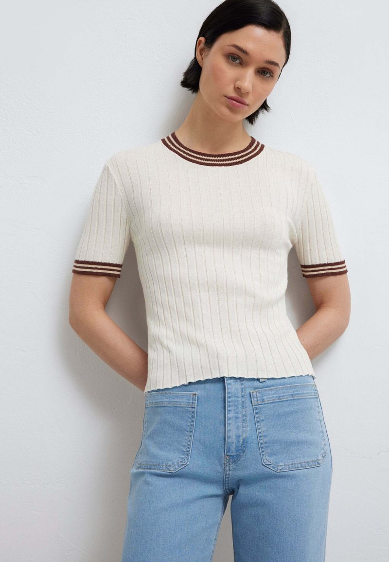 Frau mit kurzen dunklen Haaren, die einen weiß gerippten Kurzarm-Pullover mit brauner Einfassung und eine hoch taillierte hellblaue Jeans trägt, steht an einer weißen Wand.
