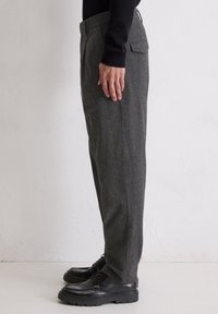Zijaanzicht van een persoon in een grijze op maat gemaakte broek, zwart lange mouwen top en zwarte veterschoenen, staand tegen een witte muur.