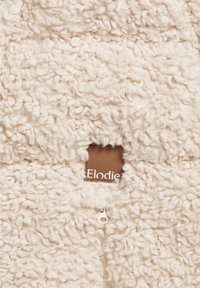Krämfärgad fleece tyg med en fluffig textur, utrustad med en liten brun etikett med texten "Elodie" och ett dragkedjestängning.