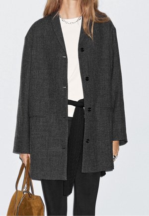 Femme portant un manteau oversize gris foncé boutonné par-dessus une chemise blanche et un pantalon noir, tenant un sac à main en daim marron dans sa main gauche.