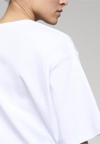 Primer plano de una persona que lleva una camiseta blanca lisa de manga corta, mostrando el cuello, el hombro y parte del rostro de perfil.