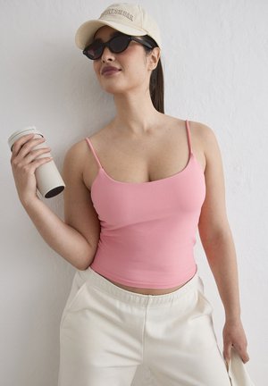 Kvinde iført pink tanktop, hvide bukser, beige kasket og sorte solbriller holder en hvid genanvendelig kop mod en hvid væg.