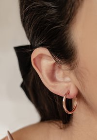 Roségoldene Creolen am Ohrläppchen, kreisförmig, mit glatter Oberfläche, glänzendem Finish und einem kleinen Scharnier für einfaches Tragen. Haare sind teilweise sichtbar.