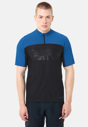 Homme portant un maillot de cyclisme bleu et noir à manches courtes avec une fermeture éclair frontale, debout devant un fond clair.
