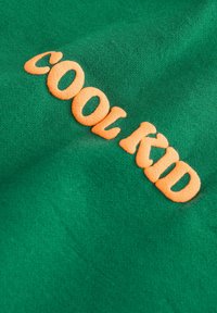 Vihreä kangas, jossa on oranssilla brodeerattu teksti 'COOL KID'. Tekstuuri on sileä, jossa on hieman kiiltoa ja rento, epämuodollinen ilme.