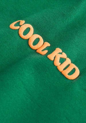 Green fabric featuring orange embroidered text "COOL KID." The texture is smooth with a slight sheen and a relaxed, casual appearance.