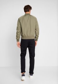 Giubbotto bomber verde oliva con polsini e orlo elasticizzati, abbinato a pantaloni neri e sneaker bianche. Design semplice, senza motivi visibili.