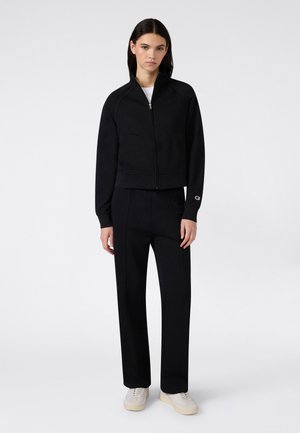 Femme debout portant une veste noire zippée, un pantalon large noir, des baskets blanches et un sous-shirt blanc sur un fond uni.