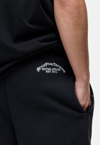Pantaloni sportivi neri con una tasca laterale, caratterizzati da testo ricamato bianco "GOOD FOR NOTHING BRITISH ATELIER EST 2011" vicino alla tasca.