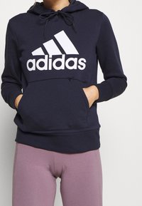 Granatowa bluza z kapturem z dużym białym logo Adidas, z regulowanym kapturem i przednią kieszenią. Wykonana z miękkiego materiału o dopasowanym kroju.