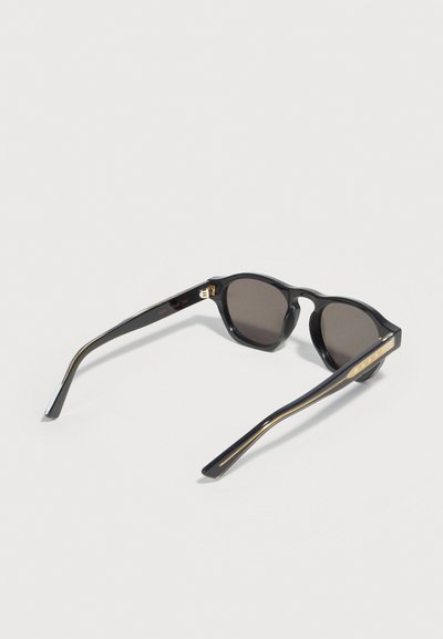 Schwarze Sonnenbrille mit dunklen runden Gläsern und goldenen Akzenten an den Bügeln, offen auf einer weißen Oberfläche liegend.