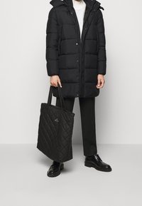 Sort pufferjakke med hætte, lynlås og trykknapper; parret med en sort quiltet totebag med et lille logo. Sorte sko fuldender looket.