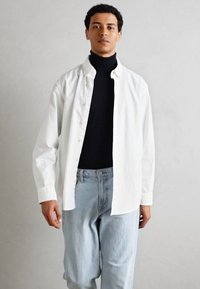 Bijela košulja s dugim rukavima preko crne turtleneck majice, uparena s laganim plavim trapericama širokog kroja. Tkanina izgleda glatko i fleksibilno.