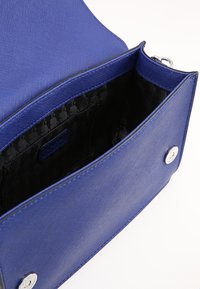 Modrá texturovaná kabelka na vybrání, otevřená, aby odhalila černou vzorovanou látkovou podšívku a stříbrné magnetické uzávěry na bílém pozadí.