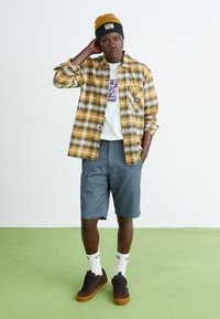 Cămașă flanelată în carouri galbene și negre peste un tricou grafic alb, pantaloni scurți gri, teniși maronii și o căciulă galbenă cu negru; șosete albe.