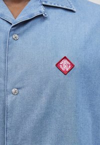 Φωτεινό μπλε πουκάμισο με ασημένια κουμπιά που φέρουν χαραγμένα αρχικά και ένα κόκκινο κεντητό σήμα σε σχήμα διαμαντιού στο στήθος.